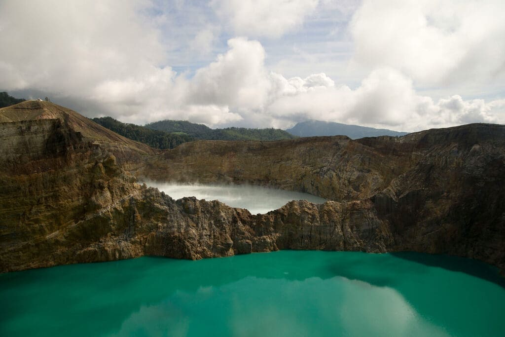 Stunning view of the turquoise crater lake at Kelimutu National Park in Flores, Indonesia, surrounded by rugged volcanic cliffs and lush forested hills under a partly cloudy sky.
