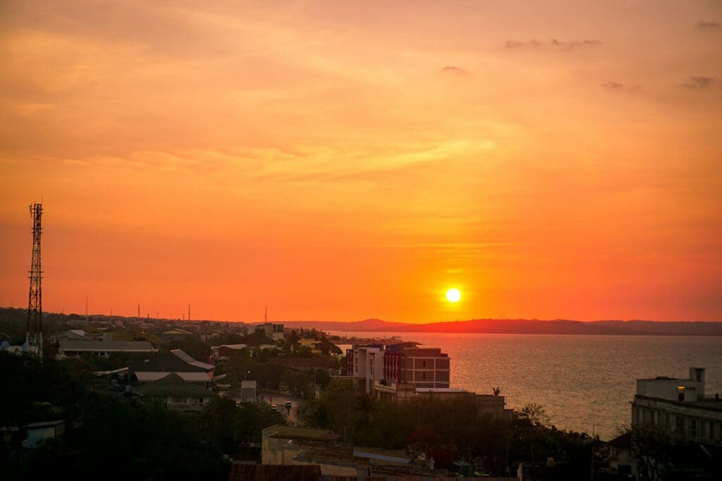 Vivid orange sunset over Kupang, Timor, Indonesia, showcasing the city's rooftops and coastline with the sun setting over the calm sea.